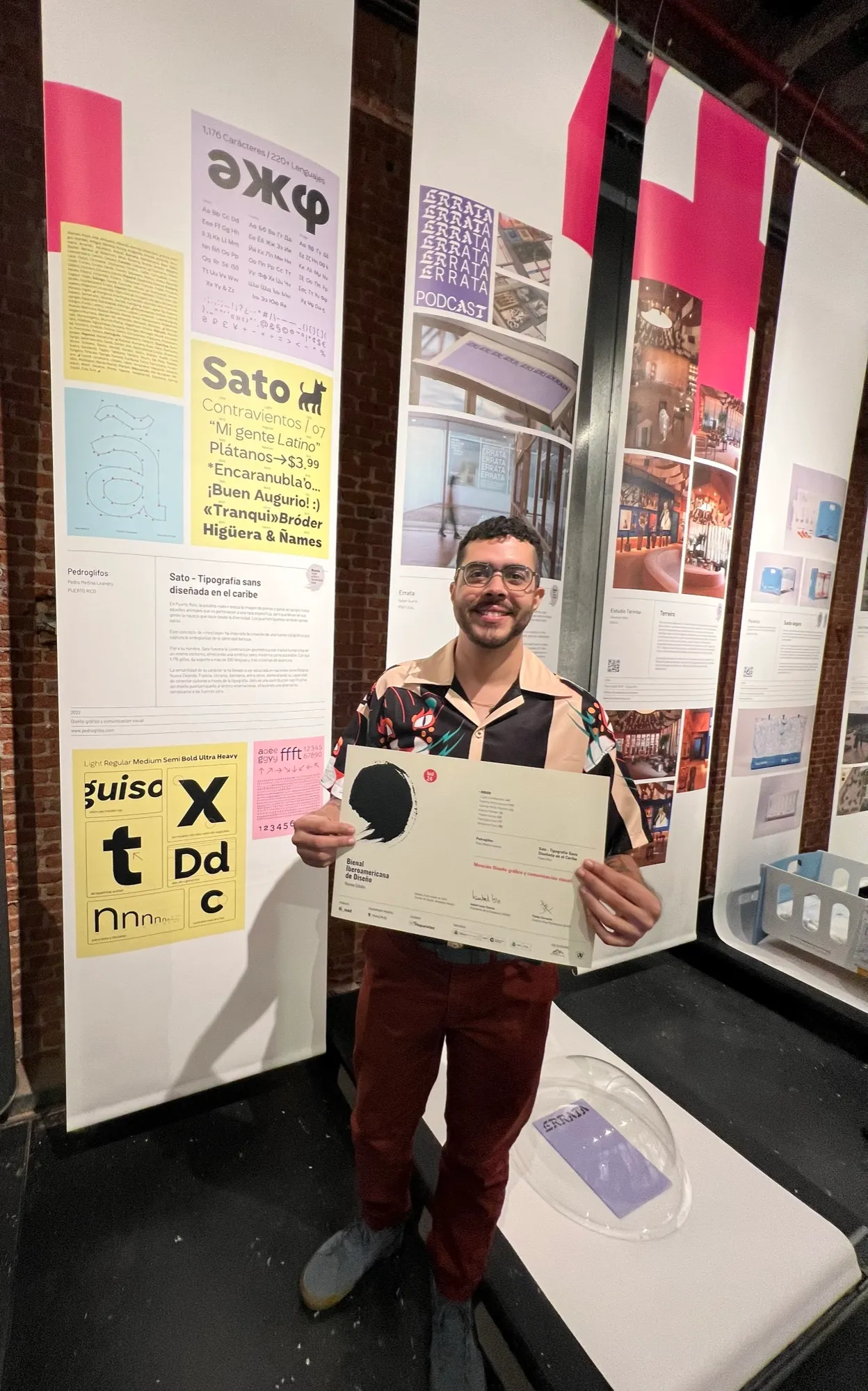 Pedro holding a Bienal Iberoamericana de Diseño certificate in front of the exhibition panel for Sato