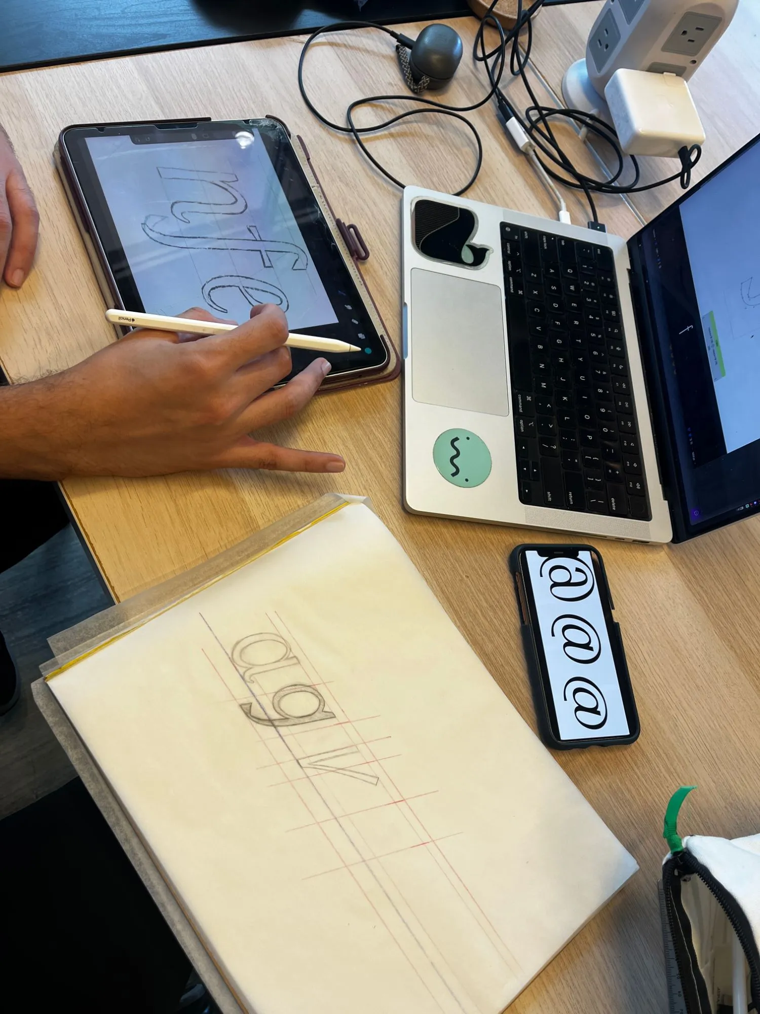 Italic letters for Modestia sketched on an iPad and on tracing paper over guidelines, beside a laptop and a phone showing @ signs
